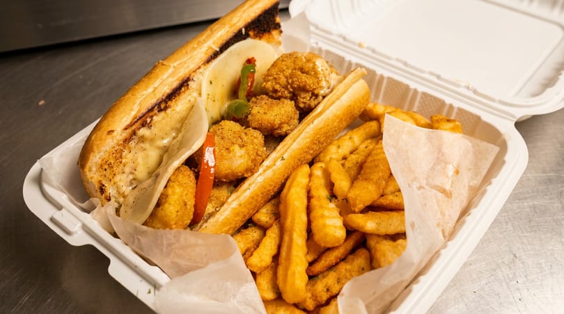 Fried shrimp po'boy sandwich with cheese, peppers, and crinkle-cut fries in a white takeout container.